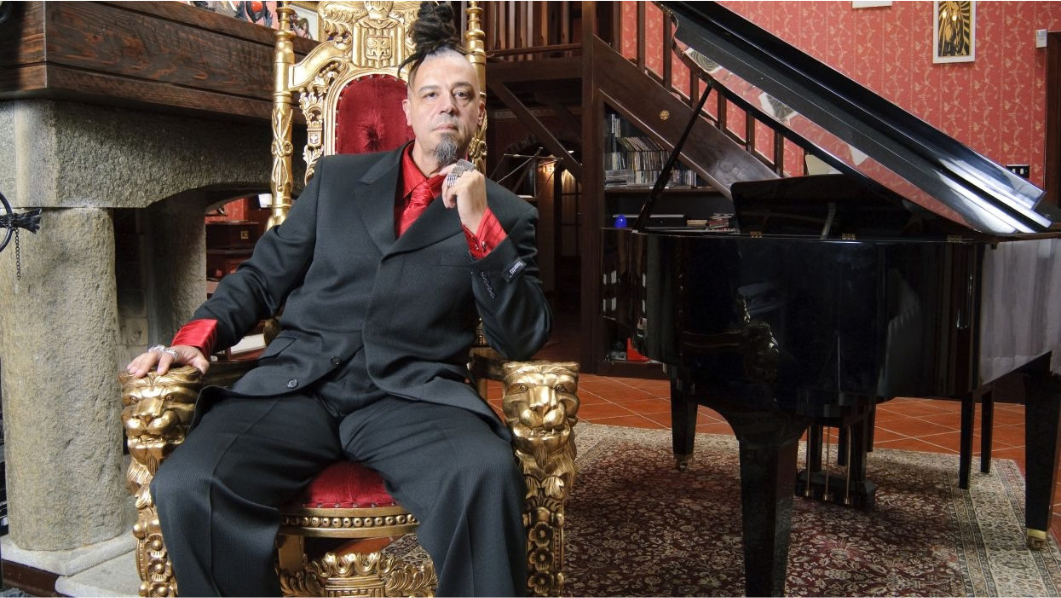 foto di Madaski seduto su una poltrona di fianco ad un pianoforte a coda