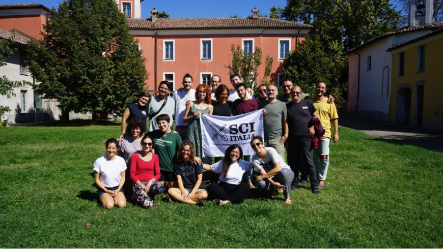 Un gruppo di persone posa sorridente all'aperto su un prato verde, tenendo una bandiera bianca con la scritta SCI ITALIA davanti a un edificio storico.