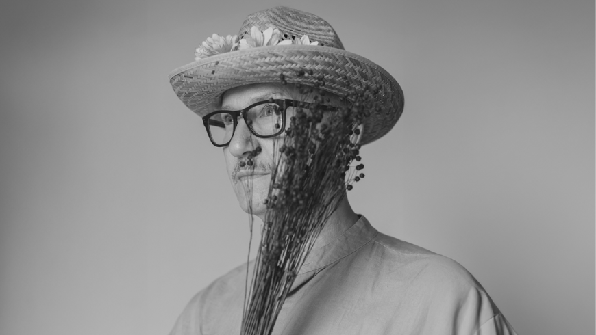 foto dell'artista che indossa un cappello di paglia e tiene davanti al viso un mazzo di fiori