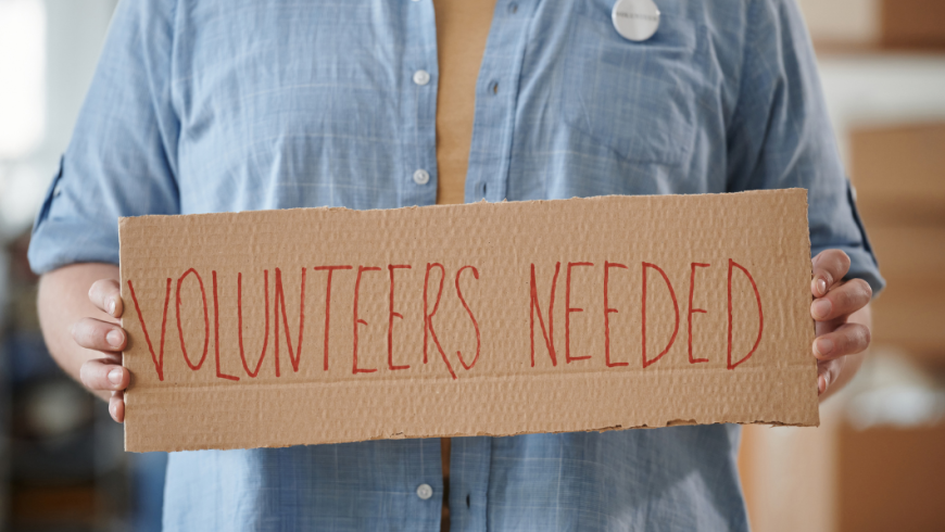 Un uomo in camicia azzurra tiene tra le mani un cartone con la scritta rossa "VOLUNTEERS NEEDED".