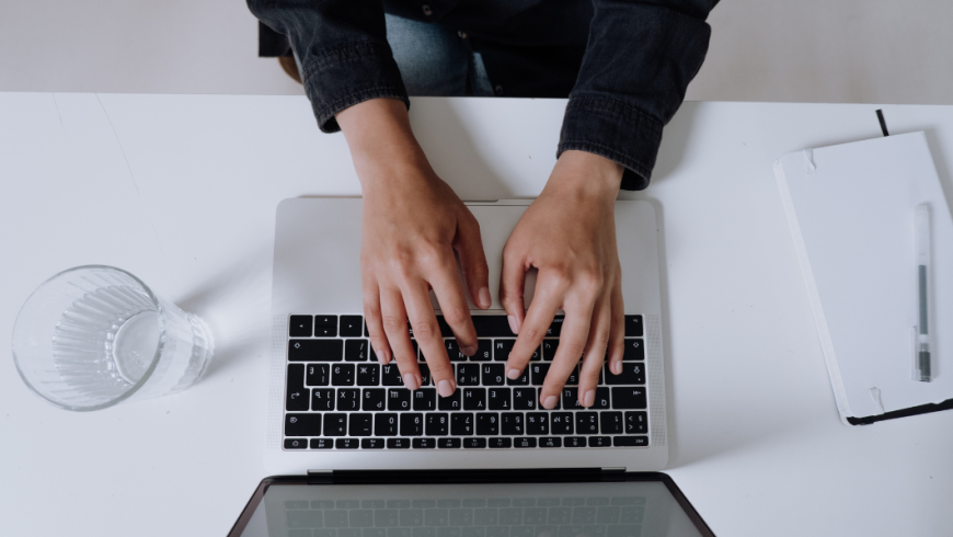 Una persona vista dall'alto digita sulla tastiera di un computer portatile posizionato su una scrivania bianca