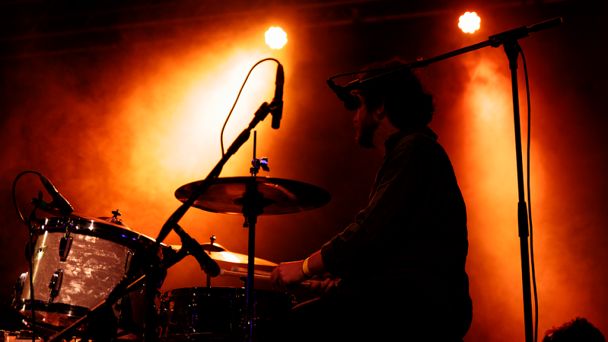 L'immagine mostra un batterista ripreso durante un'esibizione dal vivo su un palco. La scena è illuminata da luci calde di colore arancione, che creano un'atmosfera intensa e avvolgente, accentuata dalla presenza di fumo o nebbia scenica che diffonde la luce.  Il batterista è in controluce, quindi appare come una silhouette scura, con pochi dettagli visibili, ma si nota chiaramente che sta suonando la batteria, impugnando le bacchette. Indossa un braccialetto giallo al polso destro.