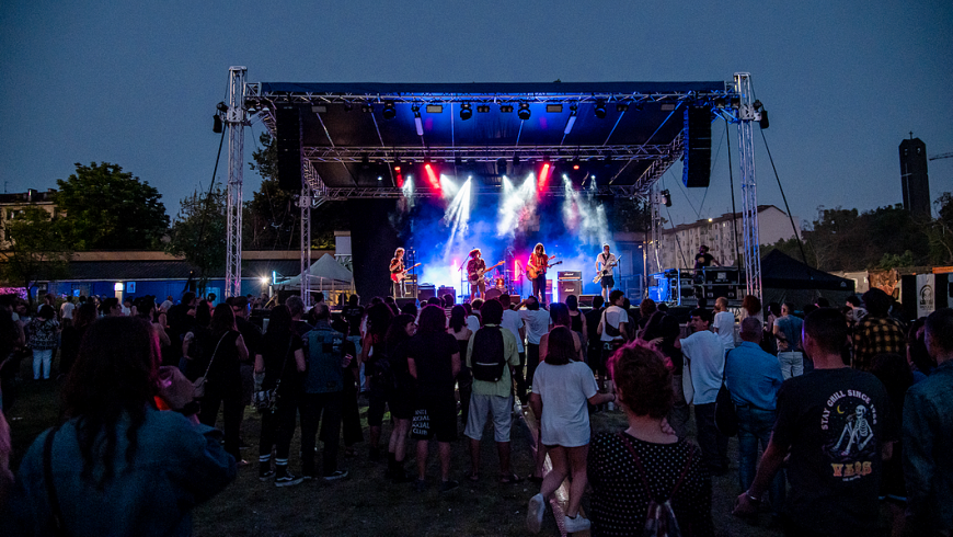 L'immagine mostra un concerto all'aperto durante il crepuscolo. Sul palco, illuminato da luci rosse e blu e avvolto da fumo, ci sono quattro musicisti che suonano chitarre. Il palco è rialzato e supportato da una struttura metallica con luci e impianti audio. Davanti al palco, una folla di persone di diverse età e stili osserva lo spettacolo. Sullo sfondo si vedono alcuni edifici, alberi e una torre alta e scura. Il cielo è di un blu profondo, tipico dell'ora serale, creando un'atmosfera suggestiva e vivace